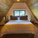 Cama doble con edredón blanco, ventana al fondo y dos mesas de noche con lámparas en un altillo de madera